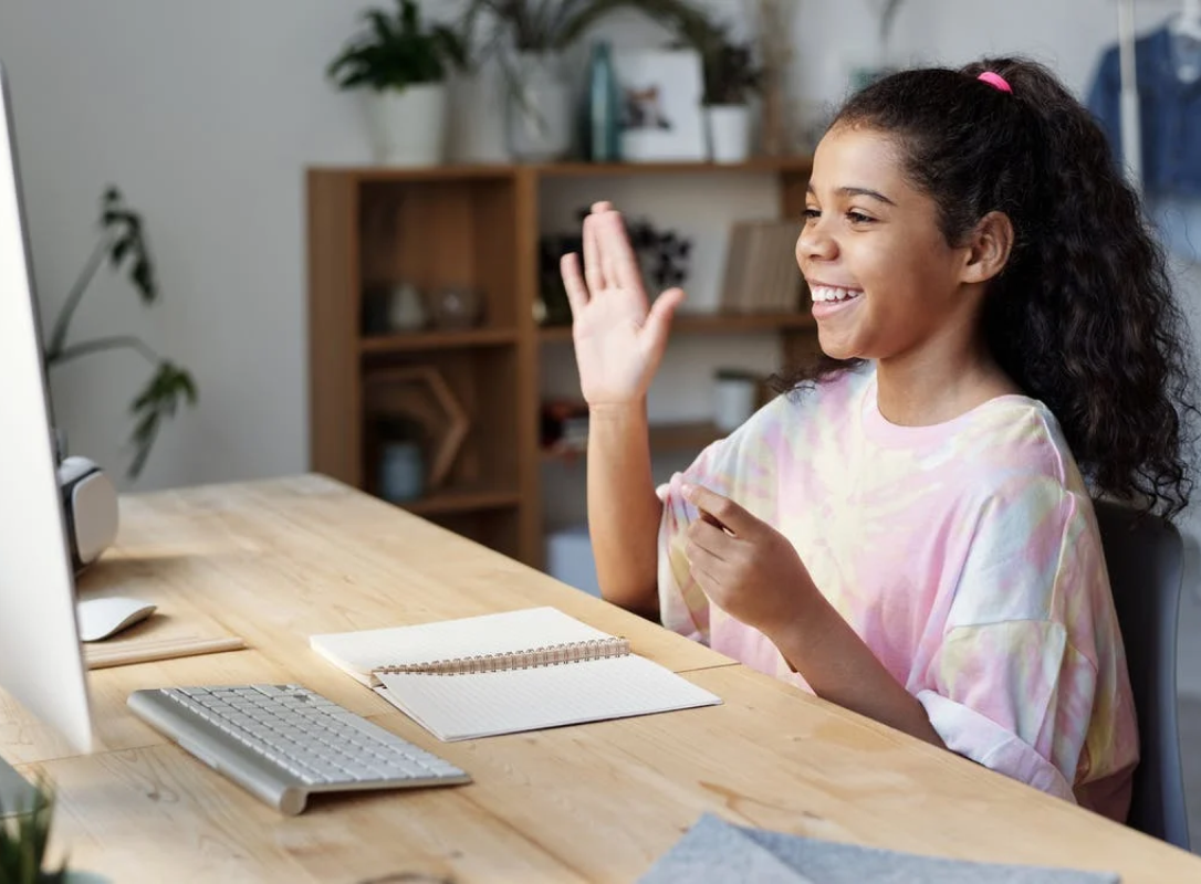 A girl taking the online class in front of a computer and raising her hand smiling
